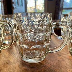 Op de afbeelding: Meerdere bierpullen van helder glas met getextureerde zijkanten en handvatten. De pullen zijn gerangschikt op een houten oppervlak. Het glas reflecteert licht, waardoor highlights en schaduwen ontstaan. De pullen zijn ontworpen voor het serveren van bier.