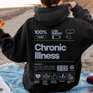 Könnte beinhalten: Schwarzer Hoodie mit einem weißen Grafikdesign, auf dem "100% Müde" und "Niedrige Energie" mit einem gebrochenen Herzen und einem Stethoskop steht. Die Grafik enthält auch die Worte "Chronische Krankheit" und eine Zutatenliste: "100% Widerstandsfähigkeit, Stärke, Anpassungsfähigkeit, ständige Schmerzen, ein bisschen medizinisches Geheimnis und nur eine Prise spontaner Nickerchen".
