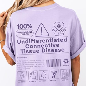 May include: A light purple t-shirt with the text "100% Autoimmune Chaos" and "Undifferentiated Connective Tissue Disease." The back of the shirt features a warning symbol, a heart with hands, a barcode, and icons representing symptoms.