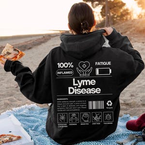 May include: A black hoodie with the words "Lyme Disease" in white. The hoodie has graphics and text indicating symptoms like fatigue, headaches, and joint pain. The person is holding a slice of pizza.