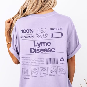 Puede incluir: Camiseta de color lila claro con la frase "Lyme Disease" en un diseño rectangular. El diseño incluye las palabras "100% Inflamado" y "Fatiga", junto con iconos y texto que describen los síntomas.