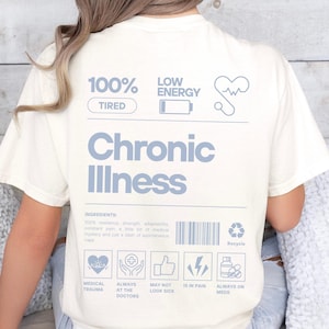 Op de afbeelding: Een wit T-shirt met een blauwe grafiek die "100% Moe" en "Lage Energie" zegt met een batterijpictogram. De grafiek zegt ook "Chronische Ziekte" en bevat een lijst met "ingrediënten", waaronder "Medische Trauma", "Altijd bij de dokter", "Ziet er misschien niet ziek uit", "Heeft pijn" en "Altijd medicijnen".