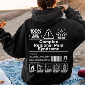 May include: Black hoodie with white text reading "100% Pain Chaos" and "Complex Regional Pain Syndrome". The back displays a warning symbol, a heart, and symptom icons. The person is holding a slice of pizza on a beach.