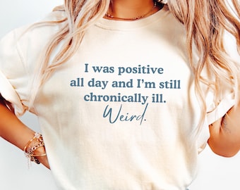 Może przedstawiać: Biały t-shirt z nadrukiem "I was positive all day and I'm still chronically ill. Weird." w kolorze niebieskim na przodzie. Koszulkę nosi osoba z długimi blond włosami i niebieskimi dżinsami.