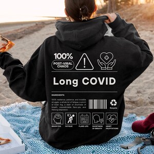 May include: Black hoodie with the text "Long COVID" and the words "100% Post-Viral Chaos." The hoodie has a warning symbol, a heart symbol, and icons representing symptoms like brain fog and fatigue. A slice of pizza is held in the left hand.