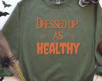 Puede incluir: Sudadera verde oliva con el texto naranja "Dressed Up As Healthy". La sudadera tiene cuello redondo y mangas largas. Decoraciones de Halloween: calavera, arañas y murciélagos.