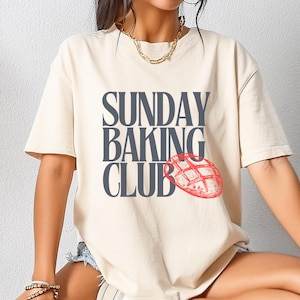 Puede incluir: Una camiseta de color crema con el texto "SUNDAY BAKING CLUB" en letras grises oscuras y una ilustración roja de una hogaza de pan.