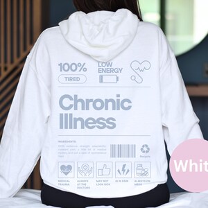May include: White hoodie with the text "100% Tired" and "Low Energy" on the front. The text "Chronic Illness" is printed in large letters below the text. The hoodie has a graphic of a heart with a heartbeat line, a battery icon, and a recycling symbol. The text "Medical Trauma", "Always at the Doctors", "May Not Look Sick", "Is in Pain", and "Always on Meds" is printed below the graphic.