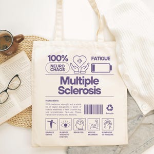 May include: Cream-colored tote bag with the words "Multiple Sclerosis" in purple. The bag features graphics and text including "100% Neuro Chaos", "Fatigue", and icons representing symptoms. A cup of coffee, glasses, and a book are in the background.
