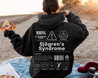 May include: Black hoodie with the text "100% Autoimmune Chaos Sjögren's Syndrome" and related graphics. The hoodie includes a list of ingredients and symptoms. The person is holding a slice of pizza.