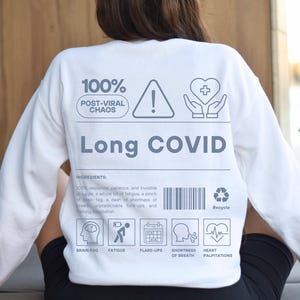 May include: White long-sleeve sweatshirt with the text "Long COVID" in gray. The back of the shirt features a graphic design with the words "100% Post-Viral Chaos" and icons representing symptoms like brain fog and fatigue.