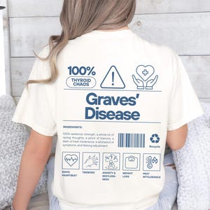 May include: Cream-colored t-shirt with blue text and graphics. The shirt features the words "Graves' Disease" and a list of symptoms, including rapid heartbeat and tremors. The top also has the text "100% Thyroid Chaos."