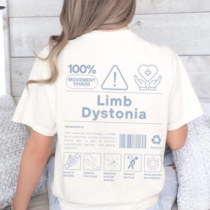 Puede incluir: Camiseta color crema con la frase "Limb Dystonia" en azul claro. La camiseta presenta un diseño gráfico con iconos y texto que incluye "100% Movement Chaos", un signo de exclamación, un corazón con una cruz y un símbolo de reciclaje.