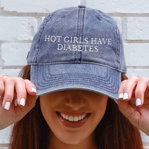 May include: A denim blue baseball cap with the embroidered text "HOT GIRLS HAVE DIABETES" in white. The cap is worn low, partially obscuring the wearer's face. The background is a white brick wall.