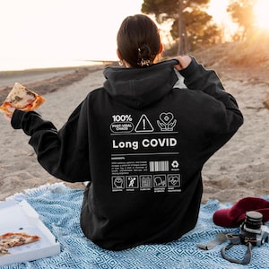 May include: Black hoodie with the text "Long COVID" and the phrase "100% Post-Viral Chaos." The back of the hoodie features a list of symptoms and a barcode. The person is holding a slice of pizza.