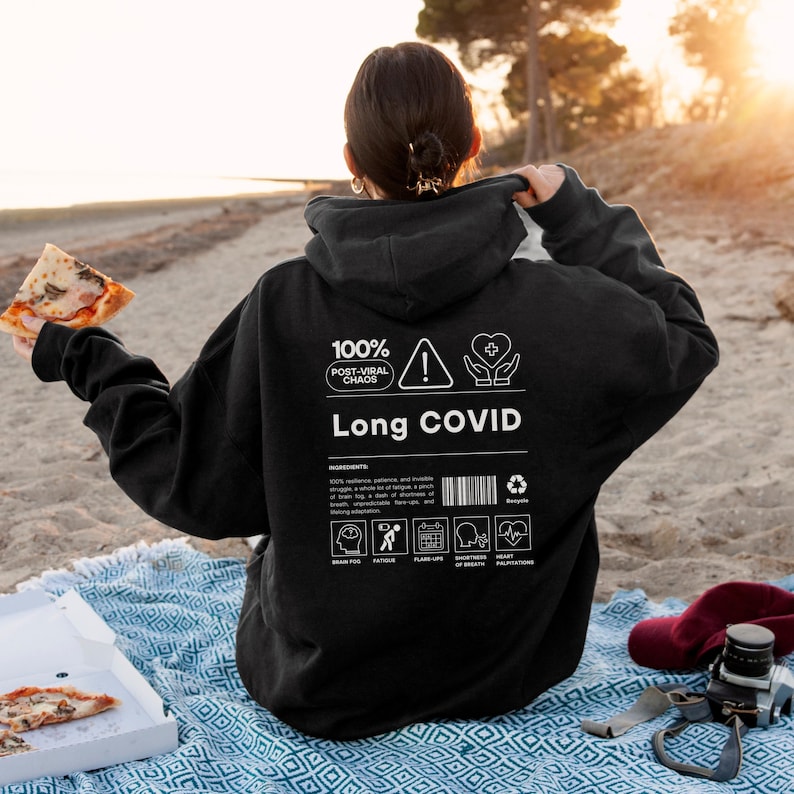 May include: Black hoodie with the text "Long COVID" and the phrase "100% Post-Viral Chaos." The back of the hoodie features a list of symptoms and a barcode. The person is holding a slice of pizza.