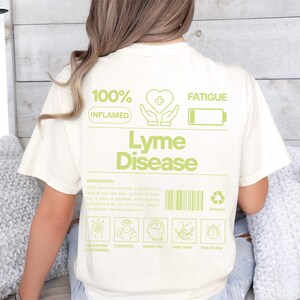 Puede incluir: Camiseta color crema con texto y gráficos verdes. El diseño incluye las palabras "Lyme Disease" y una lista de síntomas. La camiseta también presenta un gráfico de un corazón, manos y una batería.