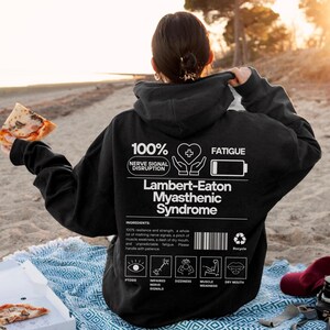 May include: Black hoodie with white text and graphics. The back of the hoodie features the text "100% Nerve Signal Disruption" and "Lambert-Eaton Myasthenic Syndrome." Additional graphics include a heart, battery, and icons representing symptoms.