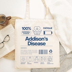 May include: Cream-colored tote bag with the words "Addison's Disease" in navy blue. The bag features icons and text related to the condition, including "100% Adrenal Chaos" and "Fatigue". A coffee mug, book, and glasses are in the background.