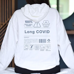 May include: White hoodie with the words "Long COVID" and other text and icons. The hoodie has a drawstring hood and long sleeves. The back of the hoodie features a list of symptoms and a recycle symbol.