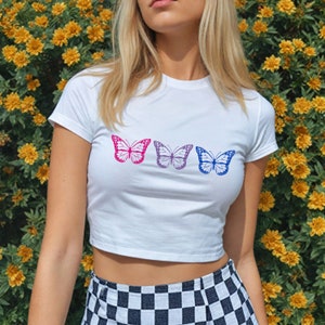 Puede incluir: Camiseta blanca recortada con una fila de tres mariposas de colores. Las mariposas son rosa, morado y azul.
