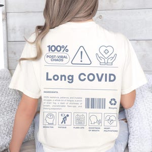 May include: Cream-colored t-shirt with the words "Long COVID" in a dark blue font. The shirt features graphics and text including "100% Post-Viral Chaos," an exclamation point, a heart with a plus sign, and icons representing symptoms like brain fog and fatigue.