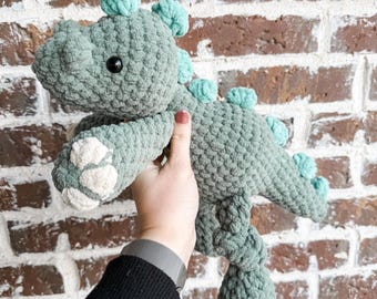 May include: A crocheted green dinosaur toy with light blue accents. The dinosaur has a white paw print on its foot.