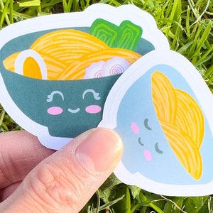 Könnte beinhalten: Zwei süße Ramen-Schüsseln mit lächelnden Gesichtern. Eine Schüssel ist blau mit gelben Nudeln und die andere ist grün mit gelben Nudeln, einem weißen Ei und grünen Zwiebeln.