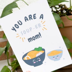 Könnte beinhalten: Eine weiße Karte mit dem Text "You are a soup-er mom!" in Blau und Orange. Die Karte zeigt zwei Zeichentrick-Schüsseln mit Ramen mit lächelnden Gesichtern.