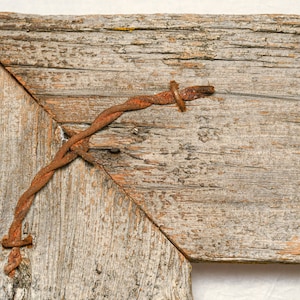 May include: A close-up of a weathered wooden frame with a rusty wire wrapped around the corner. The wood has a natural, rustic look and the wire is worn and faded.