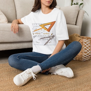 May include: A white short-sleeve t-shirt featuring a graphic print of a technical drawing on grid paper. The design includes an orange triangle ruler, a drafting compass, and the text "THEATRE LIGHTING PLOT (fig. 1)" in black ink.