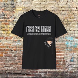 May include: A black short-sleeved t-shirt featuring the white text THEATER NINJA and MASTERS OF THE ART OF INVISIBILITY. Below the text is a cartoon ninja wearing headphones with the small white text YOU CAN'T SEE ME printed on its arm.