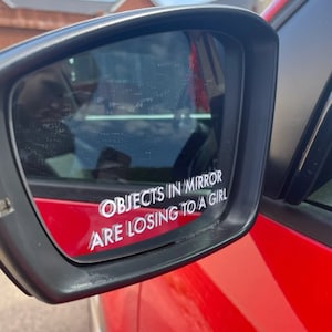 May include: A close-up of a car's side mirror with a white vinyl sticker that reads "Objects in mirror are losing to a girl".
