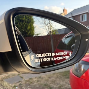 May include: A white vinyl decal applied to a car side mirror, featuring the text "OBJECTS IN MIRROR JUST GOT KA-CHOWED" in a bold, sans-serif font, flanked by two lightning bolt icons on the left and right sides of the text.