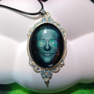 May include: Silver-toned pendant necklace featuring an oval image of a smiling face with glowing green eyes. The image is set within a decorative frame adorned with small blue gemstones and a black cord.