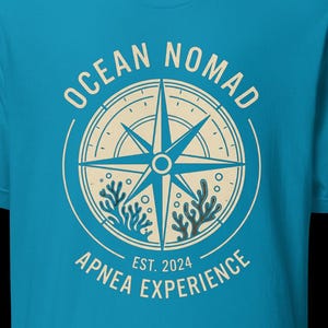 Op de afbeelding: Een felblauw t-shirt met korte mouwen en een cr&egrave;mekleurige cirkelvormige afbeelding op de borst. Het ontwerp bevat een kompasroos, koraalillustraties en de tekst "OCEAN NOMAD" en "APNEA EXPERIENCE EST. 2024" in een strak, schreefloos lettertype.