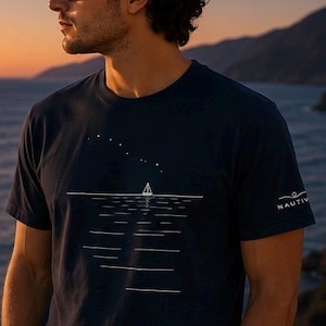 Könnte beinhalten: Ein dunkelblaues Kurzarm-T-Shirt mit einer weißen Grafik eines Segelboots auf dem Wasser unter einem Sternenhimmel. Auf dem Ärmel befindet sich der Schriftzug NAUTIV in Weiß. Das T-Shirt hat einen Rundhalsausschnitt und eine Standardpassform.