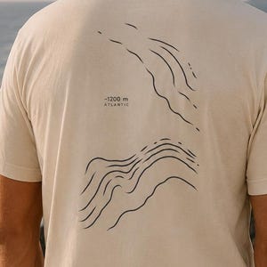 Könnte beinhalten: Ein cremefarbenes Kurzarm-T-Shirt mit einer schwarzen Linien-Grafik auf der Rückseite. Das Design zeigt abstrakte topografische Wellenmuster und enthält den aufgedruckten Text -1200 m ATLANTIC. Das T-Shirt hat einen Rundhalsausschnitt.