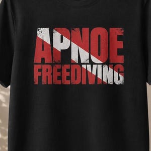 Könnte beinhalten: Ein schwarzes kurzärmeliges T-Shirt mit einem Grafikdruck auf der Brust. Das Design zeigt das Wort APNOE über dem Wort FREEDIVING in einer abgenutzten roten Schrift, überlagert von einem weißen diagonalen Streifen, der an eine Taucherflagge erinnert.