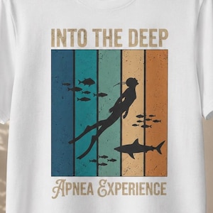 Könnte beinhalten: Ein weißes Kurzarm-T-Shirt mit einem Grafikdruck und dem Text INTO THE DEEP APNEA EXPERIENCE. Das Design zeigt die Silhouette eines Tauchers und eines Hais vor vertikalen Streifen in Blau-, Türkis- und Orangetönen sowie kleine Fische.
