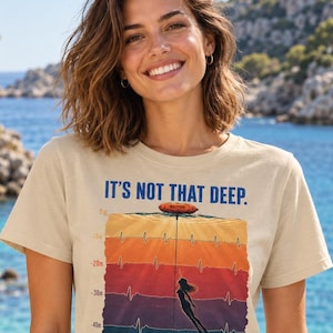 May include: A beige short-sleeve t-shirt featuring a graphic of a freediver descending through colorful water layers with depth markers from 0m to 50m, an EKG heartbeat line, and the text IT'S NOT THAT DEEP. IN THE DEEP. printed in blue.