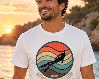 Może przedstawiać: Biały t-shirt z krótkim rękawem z okrągłą grafiką przedstawiającą zachód słońca w stylu retro, sylwetkę nurka swobodnego oraz napis "APNEA REBELLION EST. 2024 ~ONE KICK AT A TIME~". Koszulka zestawiona jest z oliwkowozielonymi szortami.
