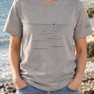 Peut inclure: Un t-shirt gris chin&eacute; &agrave; manches courtes avec un motif noir minimaliste repr&eacute;sentant une ligne d'horizon, un petit voilier et des points dispers&eacute;s au-dessus. Le mot NAUTIVE est imprim&eacute; en petites lettres noires sur la manche gauche.