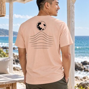 K&ouml;nnte beinhalten: Ein pfirsichfarbenes Kurzarm-T-Shirt mit einer schwarzen Grafik auf der R&uuml;ckseite. Das Design besteht aus einer kreisf&ouml;rmigen Mondform &uuml;ber einer Reihe von f&uuml;nf horizontalen, wellenf&ouml;rmigen Linien, die wie Meereswellen oder H&uuml;gel wirken.