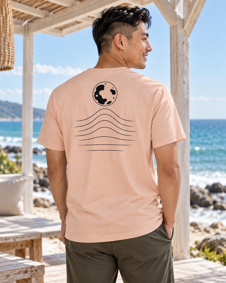 K&ouml;nnte beinhalten: Ein pfirsichfarbenes Kurzarm-T-Shirt mit einer schwarzen Grafik auf der R&uuml;ckseite. Das Design besteht aus einer kreisf&ouml;rmigen Mondform &uuml;ber einer Reihe von f&uuml;nf horizontalen, wellenf&ouml;rmigen Linien, die wie Meereswellen oder H&uuml;gel wirken.