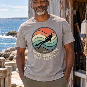 K&ouml;nnte beinhalten: Ein beiges, meliertes Kurzarm-T-Shirt mit einer kreisf&ouml;rmigen Grafik, die einen Retro-Sonnenuntergang mit orangefarbenen, gelben und t&uuml;rkisfarbenen Wellen zeigt. Eine schwarze Silhouette eines Apnoetauchers ist zentriert mit dem Text "APNEA REBELLION EST. 2024 ONE KICK AT A TIME".