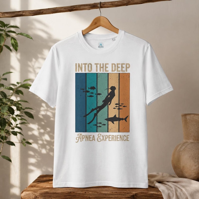 Könnte beinhalten: Ein weißes Kurzarm-T-Shirt mit einem Grafikdruck und dem Text INTO THE DEEP APNEA EXPERIENCE. Das Design zeigt die Silhouette eines Tauchers und eines Hais vor vertikalen Streifen in Blau-, Türkis- und Orangetönen sowie kleine Fische.