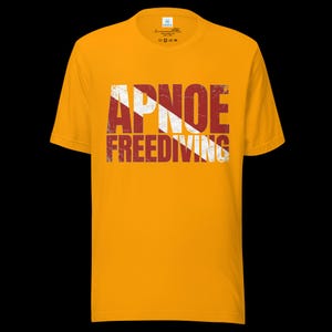 Puede incluir: Una camiseta de manga corta color amarillo mostaza con el texto APNOE FREEDIVING en una fuente desgastada y envejecida. Las letras est&aacute;n superpuestas con un patr&oacute;n de rayas diagonales blancas sobre un fondo rojo oscuro, creando un aspecto vintage.