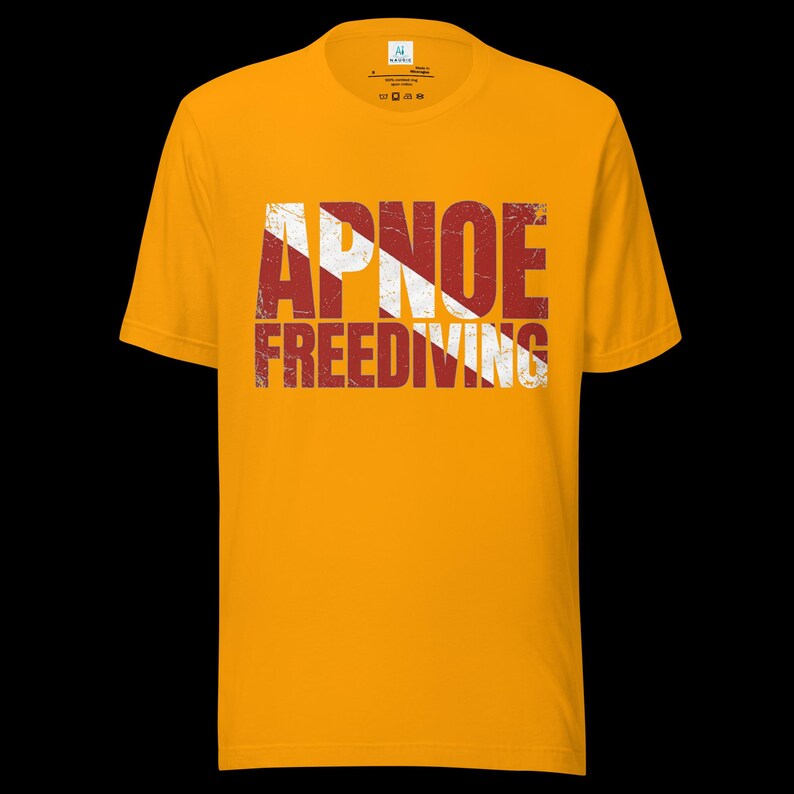Puede incluir: Una camiseta de manga corta color amarillo mostaza con el texto APNOE FREEDIVING en una fuente desgastada y envejecida. Las letras est&aacute;n superpuestas con un patr&oacute;n de rayas diagonales blancas sobre un fondo rojo oscuro, creando un aspecto vintage.