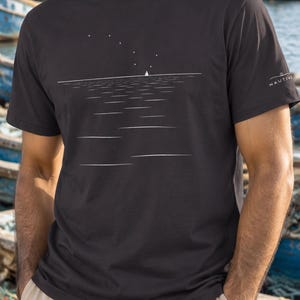 Peut inclure: Un t-shirt &agrave; manches courtes gris anthracite fonc&eacute; orn&eacute; d'un graphique lin&eacute;aire blanc minimaliste repr&eacute;sentant un voilier sur l'eau sous un ciel &eacute;toil&eacute;. La manche gauche porte le texte blanc NAUTIVE avec une petite ic&ocirc;ne de bateau.